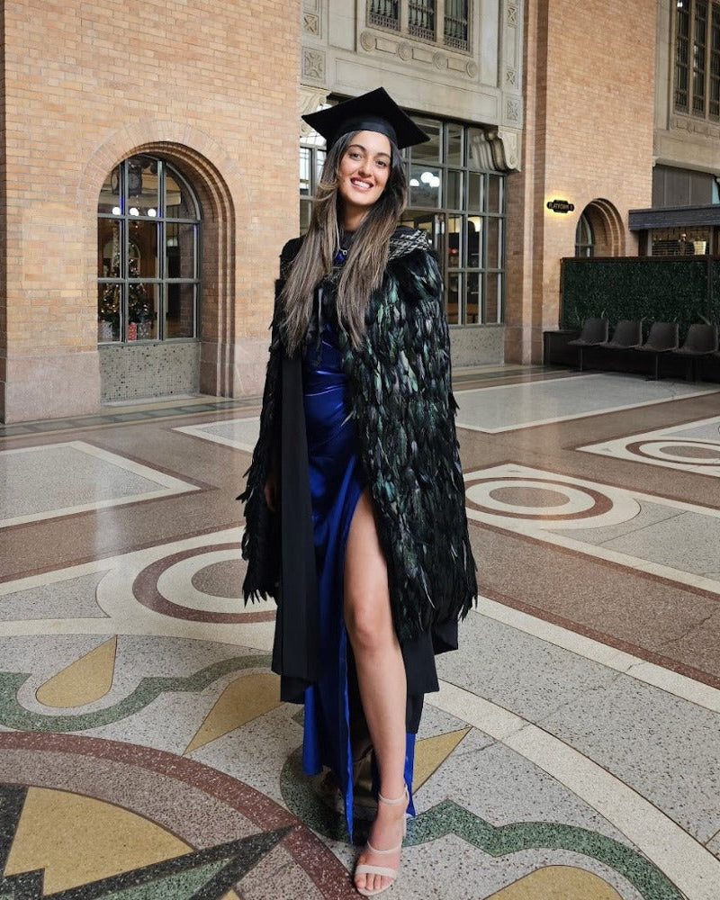 A person wearing a traditional Māori korowai with a shimmering Niho Taniwha taniko band design, standing in a large hall with decorative flooring.