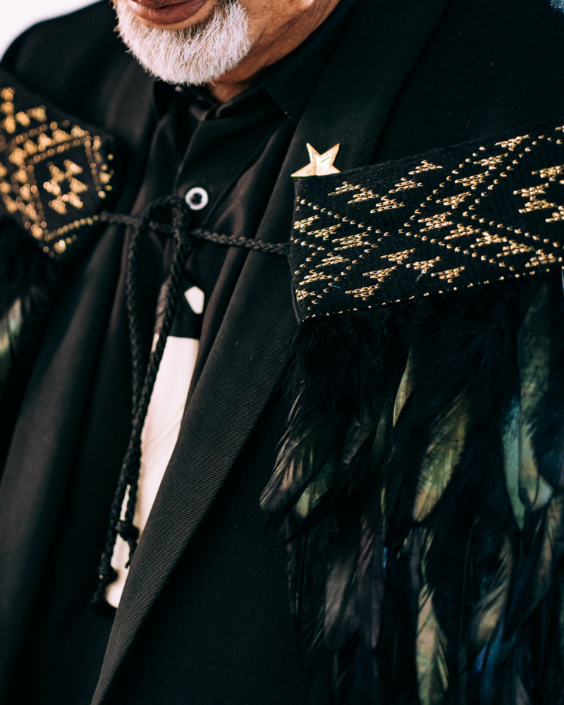 Close-up of a Māori korowai featuring a Niho Taniwha taniko band design with gold and black patterns on black feathers, worn by a person.