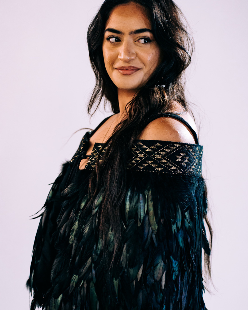 Close-up of a Māori korowai featuring a Niho Taniwha taniko band design with gold and black patterns on black feathers, worn by a young woman