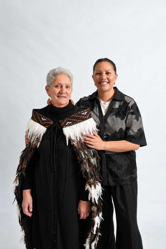 Two women smiling together, with the woman in front wearing the chocolate Tuku Iho korowai and KBH tāniko band.