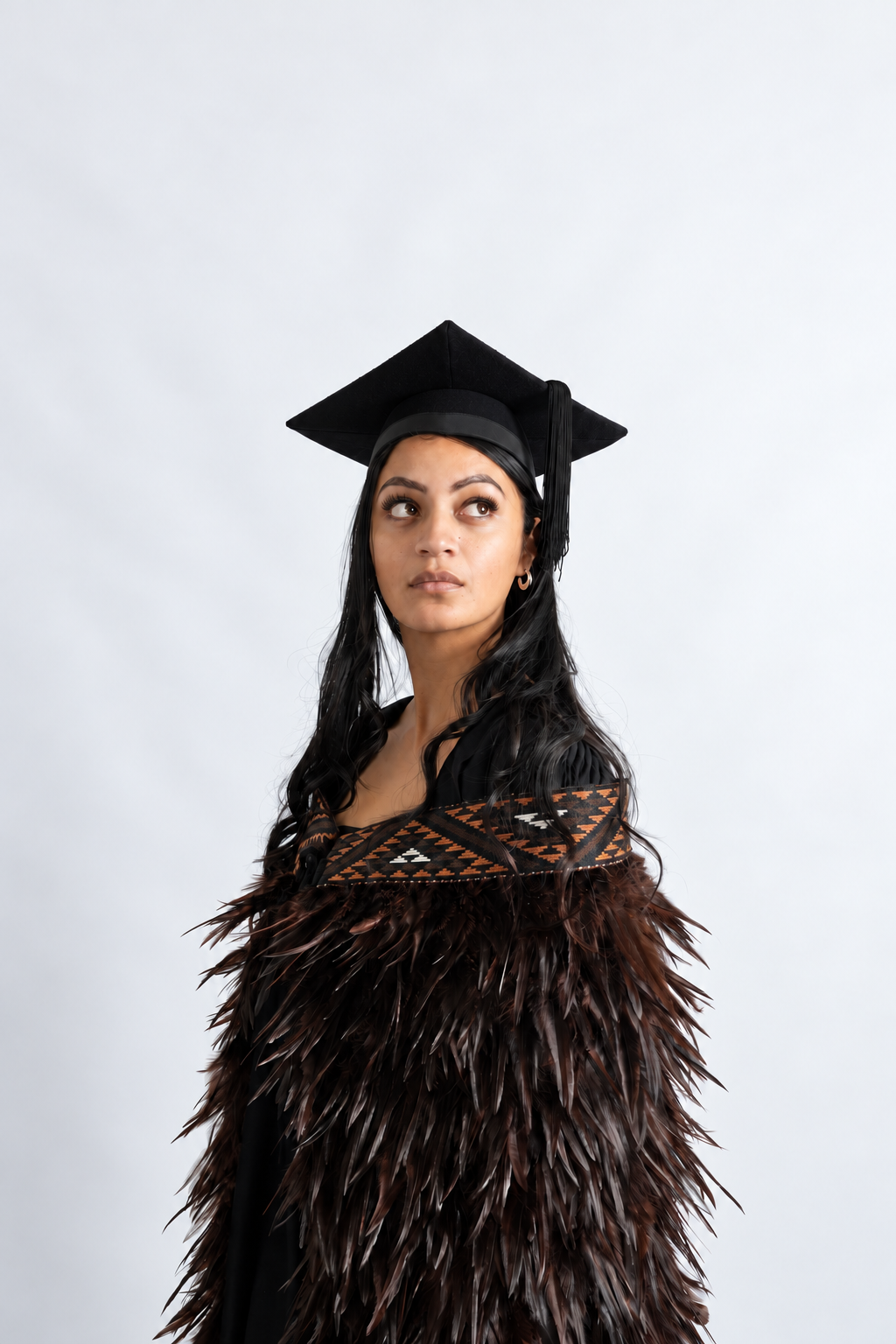 Graduate wearing the full-length Kahu Kiwi korowai with deep chocolate feathers and brown geometric tāniko band, looking upward.