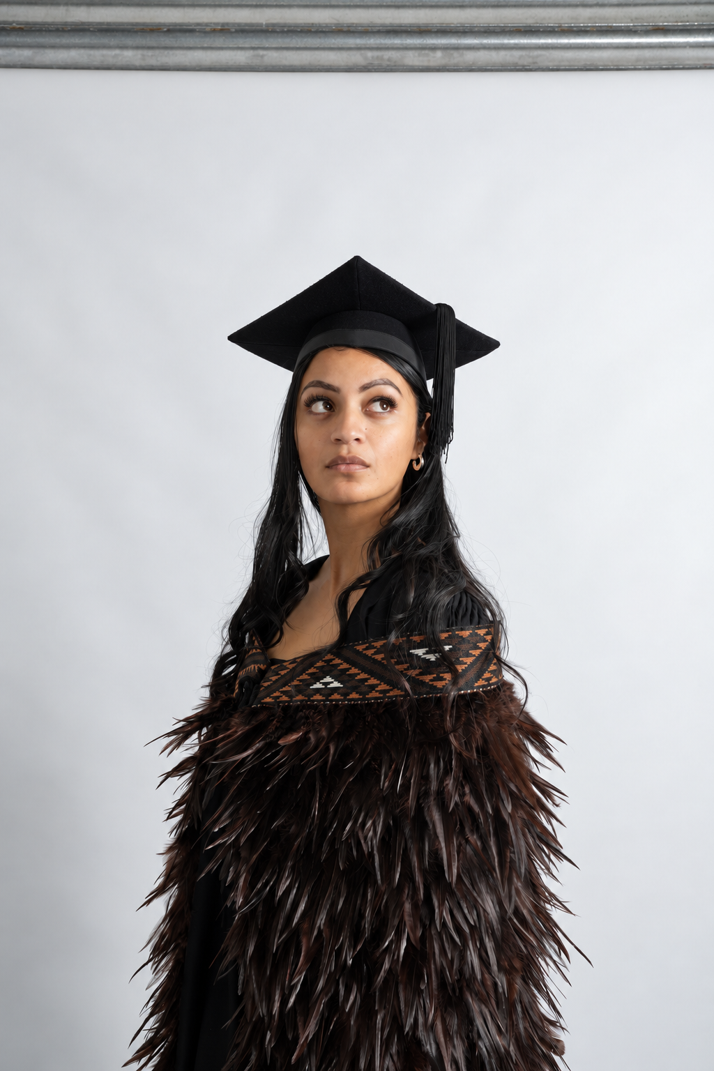 Graduate wearing the full-length Kahu Kiwi korowai with deep chocolate feathers and brown geometric tāniko band, looking upward.