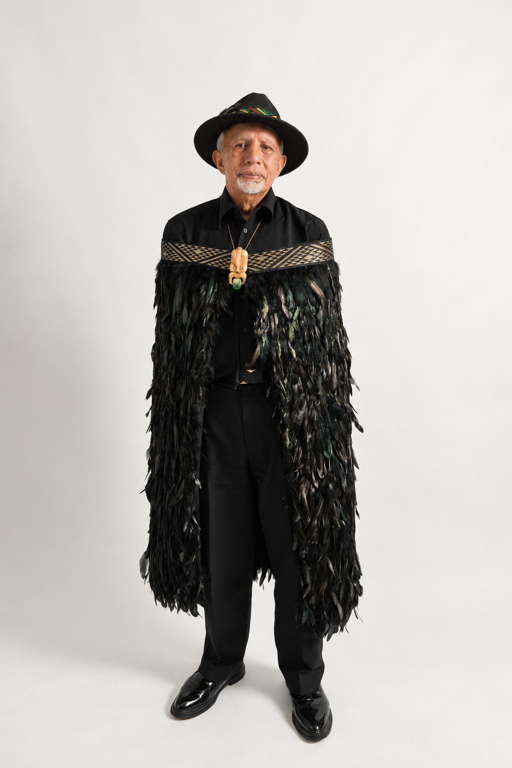 Male model wearing full-length black Pīataata korowai with a gold tāniko band, showing the complete feather drop.