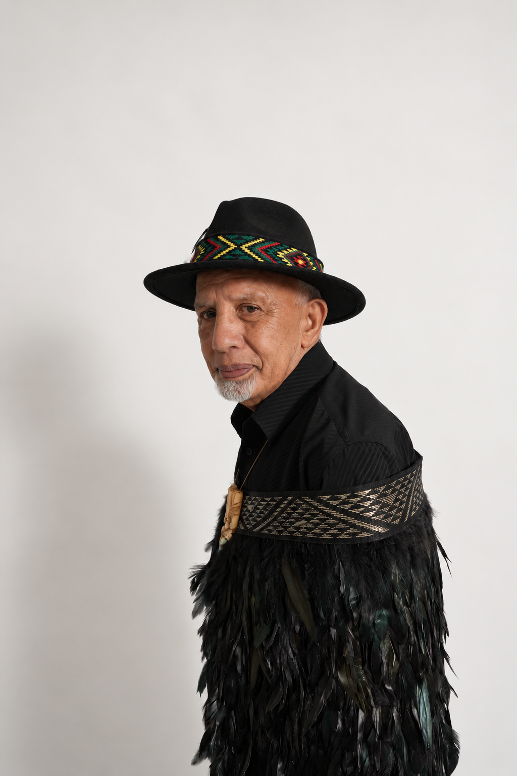 Closer side portrait of a male model wearing full-length black Pīataata korowai with a gold tāniko band.