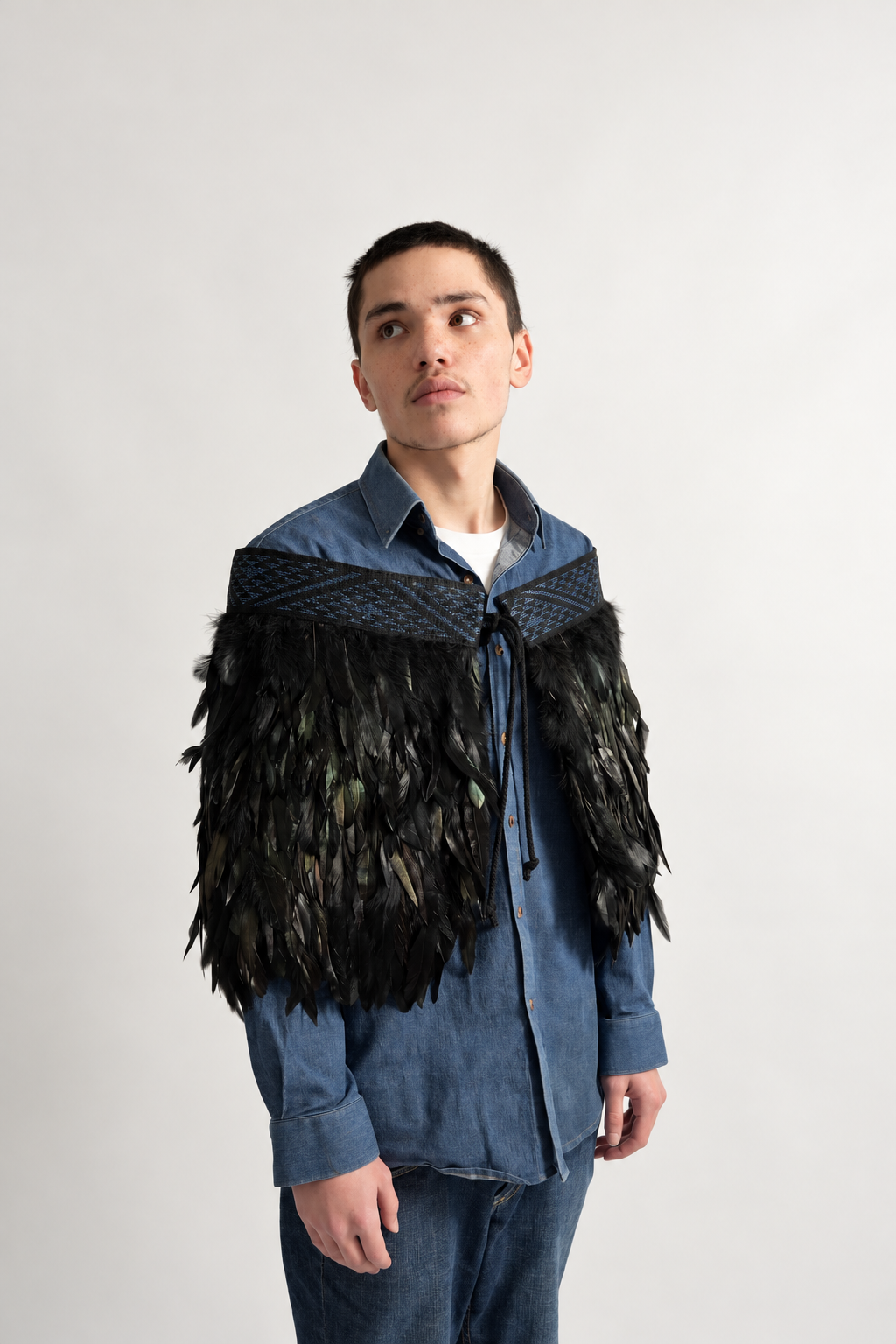Male model wearing black Pīataata korowai with a pāua blue tāniko band, looking upwards.